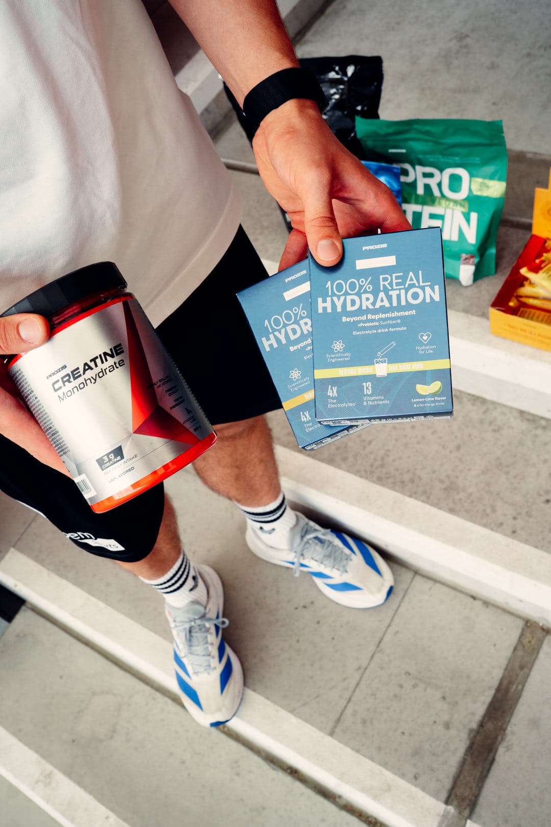 Prozis Kreatin und Hydration in den Händen, daneben das Protein auf den Stufen